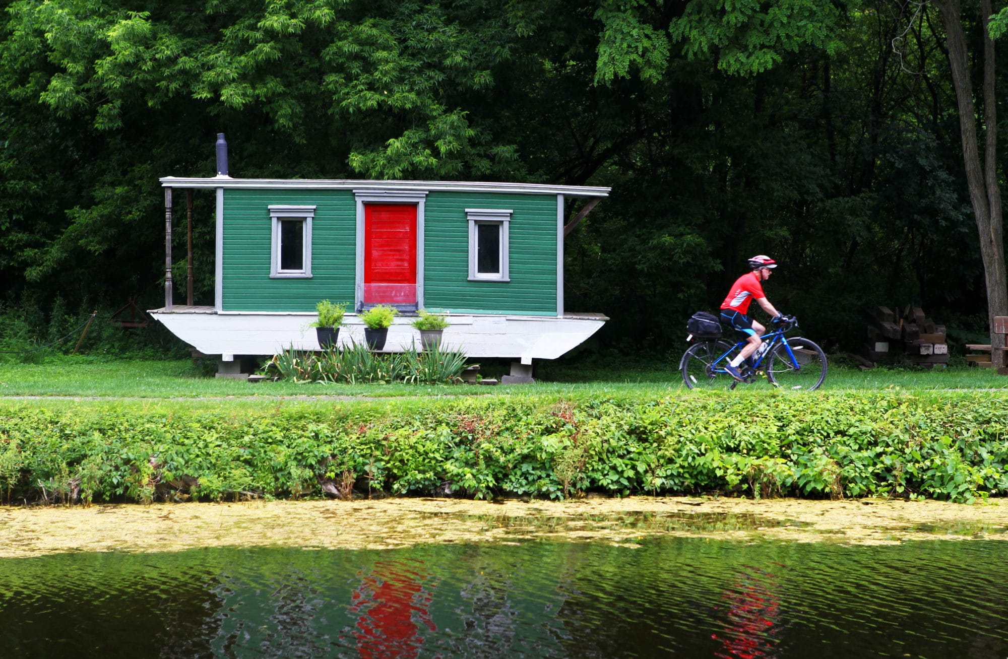 A cyclist rides past a red door of a houseboat, used as a home, in summer, along the Erie Canalway Trail, part of the longer Empire State Trail in New York State.