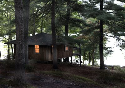 A rustic camp by the water at twilight, with a single window lit.