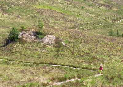 A wide, green view of the Scottish Highlands, with a hiker walking on the winding trail through bare landscape