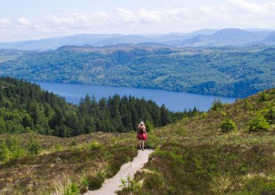 A solitary hiker on a dirt path through the scottish highlands, with a loch and mountains in the distance